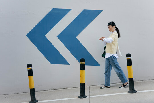 Woman walking in urban area checking watch near blue arrow sign
