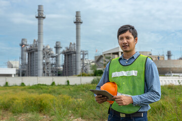 Engineer wearing safety vest holding orange helmet and tablet at industrial plant site,...