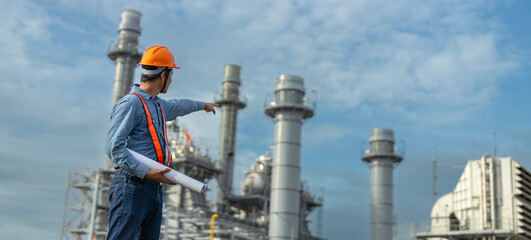 Engineer wearing safety gear pointing towards industrial power plant structures while holding...