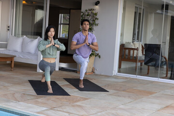 Fototapeta premium Diverse yoga partners holding forward lunge on mats at poolside terrace beside sliding doors