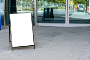 Blank sidewalk sandwich board sign placed outside a modern glass-front building. Empty white...