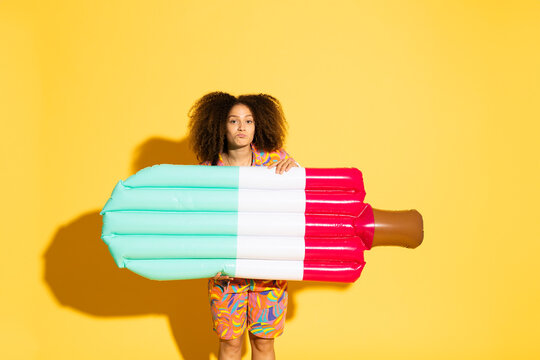 Young woman holding pool raft in front of yellow background