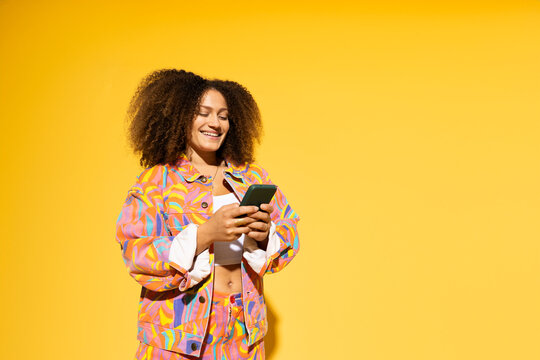 Happy woman with curly hair using smart phone against yellow background