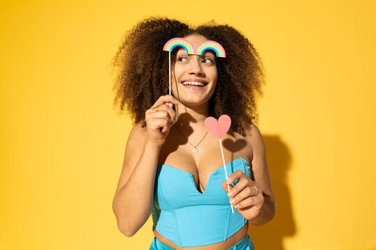 Smiling young woman with heart and rainbow prop posing against yellow background