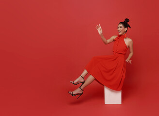Asian woman in red dress sitting on cube expressing festive confidence and celebration with elegant fashion joyful emotion cultural holiday spirit bold color and modern studio portrait.