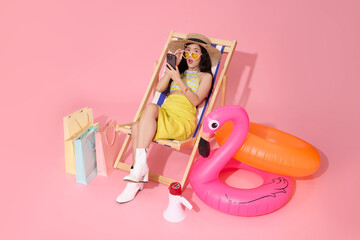 Fashionable young asian woman relaxing on deck chair with smartphone and shopping bags beside inflatable flamingo expressing summer vacation mood and digital lifestyle connection