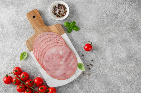 Delicious ham cut into round slices on a cutting board with spices, cherry tomatoes and basil leaves on a gray stone background. Top view.