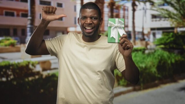 Man holding green gift with thumb pointing to self and raised hand on street near building and palm, smiling while presenting the present; joy celebration.