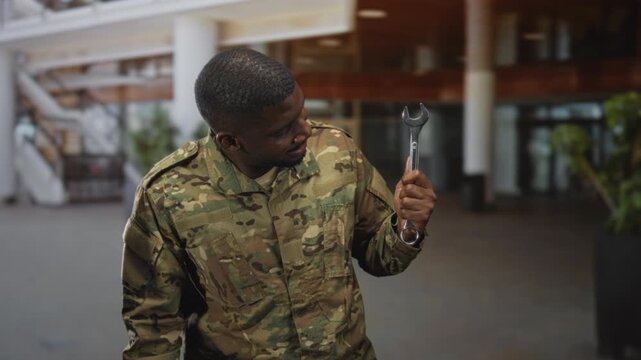 Young man soldier in camouflage uniform holding a wrench and looking at the tool while standing inside a building with columns and plants; duty focus.