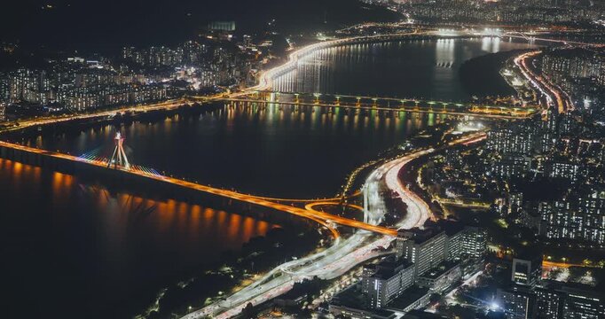 Aerial perspective captures a bustling city at night with bright lights reflecting on the river, highlighting bridges and moving vehicles. Time-lapse, Seoul, South Korea