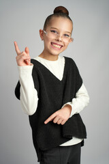Cheerful young girl wearing glasses and a black sweater vest smiles broadly while pointing her index finger upward. Bright studio portrait on a neutral grey background