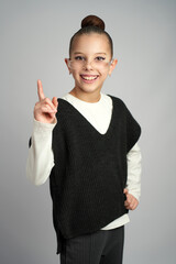 Cheerful young girl wearing glasses and a black sweater vest smiles broadly while pointing her index finger upward. Bright studio portrait on a neutral grey background