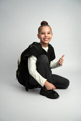 Happy Schoolgirl with Backpack Giving Thumbs Up while Sitting on Floor. Close-up studio portrait on a neutral grey background.