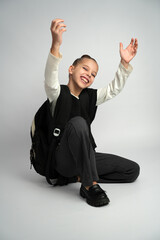 Joyful young girl in a school uniform and backpack crouching while laughing and throwing her hands up in celebration. Studio portrait on a neutral grey background