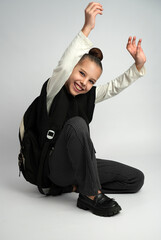 Joyful young girl in a school uniform and backpack crouching while laughing and throwing her hands up in celebration. Studio portrait on a neutral grey background