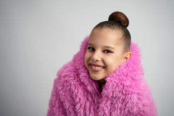 Playful portrait of a young girl with a bun hairstyle wearing a pink fur jacket. She is leaning forward with a joyful expression against a neutral grey background