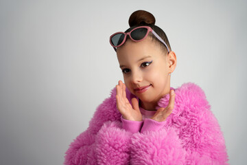 Close-up of a smiling girl in a fluffy pink jacket and sunglasses on her head, framing her face with her hands against a grey backdrop