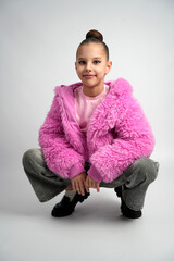 A stylish girl wearing a bright pink faux fur jacket and pink shirt crouching and smiling against a clean grey studio background