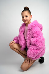 A stylish girl wearing a bright pink faux fur jacket and pink shirt crouching and smiling against a clean grey studio background