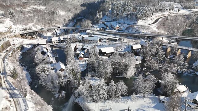 Drone follows a snow-covered road curving toward a bridge in Rastoke, Croatia. Waterfalls and river visible in a magical winter landscape with snow and soft daylight.
