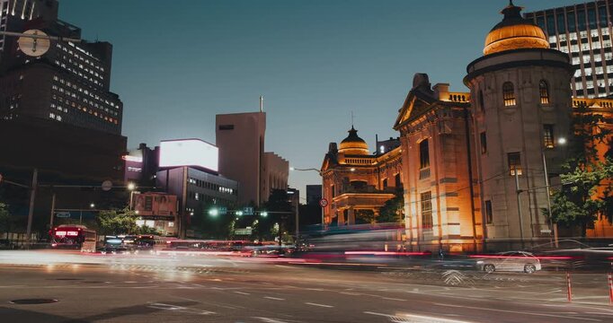 An urban traffic scene unfolds at dusk, showcasing illuminated historical buildings and moving vehicles creating light trails across the busy intersection. Time-lapse, Seoul, South Korea