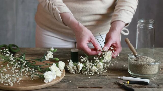 Florist at work: woman shows how to make romantic candle holder using a glass jar, carnations flowers, gypsophila paniculata twigs and sand. Step by step, tutorial. Images available.