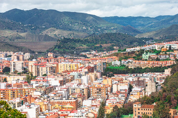 Fototapeta premium Residential districts of Malaga, Spain, with rolling Montes de Malaga mountains in background. Dense urban landscape features numerous multi-story buildings and a large dam nestled in distant hills