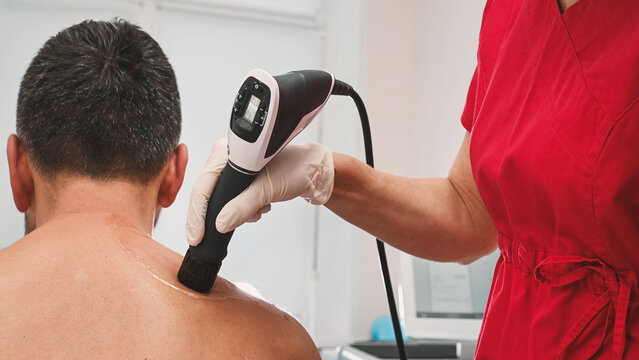 A rehabilitation doctor performs shockwave therapy procedure on a male patient, massaging his shoulder, posture check, spine health, orthopedic prevention and nervous system concept