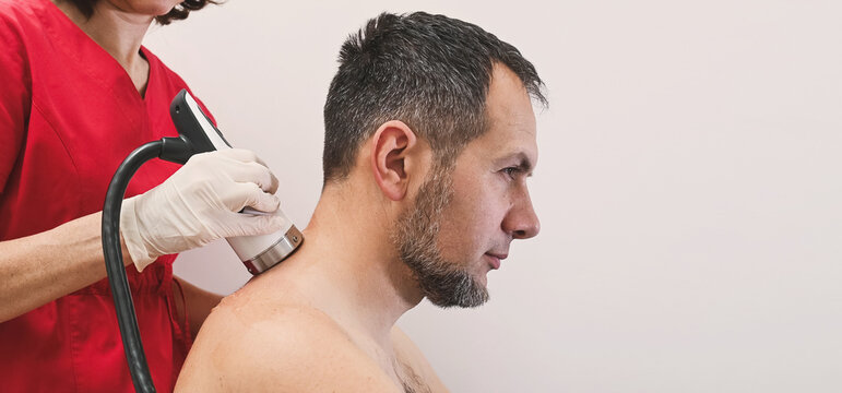 A rehabilitation doctor performs shockwave therapy procedure on a male patient, massaging his shoulder, posture check, spine health, orthopedic prevention and nervous system concept copy space banner