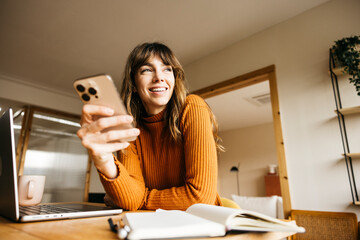 Woman smiling while holding smartphone and working on laptop at her desk © Jordi Salas