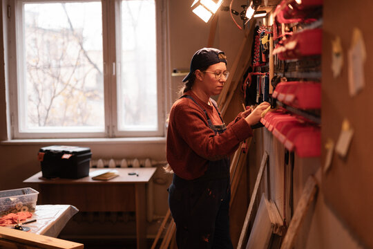 Confident lady artisan reaching for instrument from storage rack inside craft studio