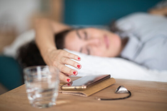 Sleepy Woman lying in bed reaching out to turn off the alarm on her mobile phone