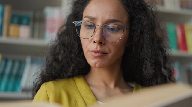 Calm Latina Hispanic woman in eyeglasses reading book in quiet library female student girl turning pages literature bookworm learner reader scholar glasses study novel exam preparation hobby academic