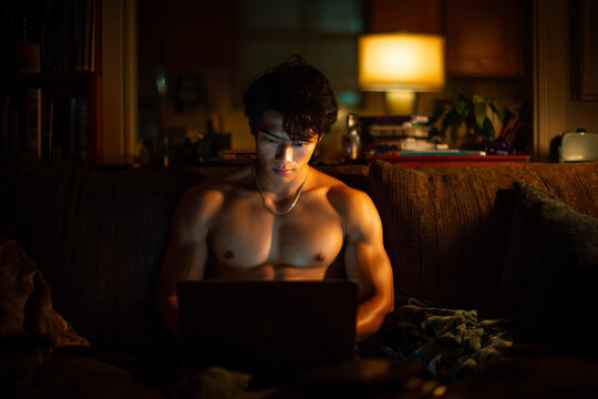 Muscular Young Man Sitting Shirtless on a Sofa Using a Laptop at Night in a Warmly Lit Living Room