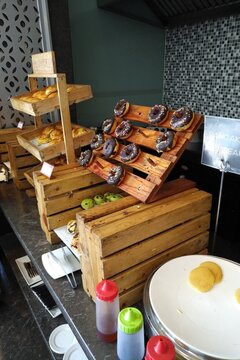 Hotel breakfast buffet with donuts, pastries, and dessert toppings arranged on rustic wooden stands. Perfect for hospitality, travel, restaurant, catering, and food service design projects.