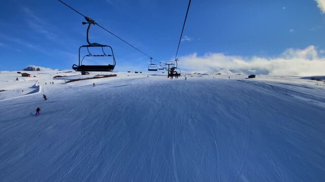 A wide-angle shot of skiers on a chairlift at Alpe di Siusi in the Dolomites. A panoramic shot of the lift ascending the snow-covered slopes of the Seiser Alm. Winter sports in the Italian Alps skiers