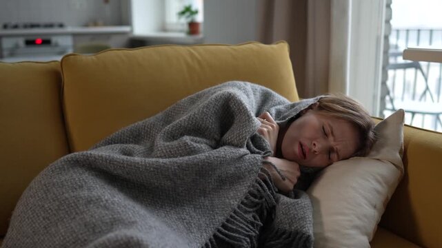Sad lost young woman feels unwell lying on sofa wrapped in wool plaid, showing first signs of cold and flu symptoms. Sick female suffering from chills, curling on couch with fever by virus infection
