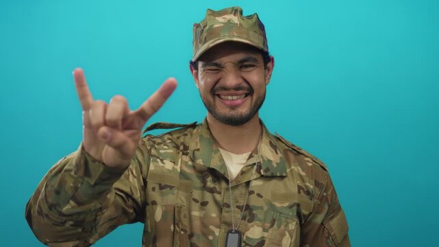 Young hispanic man in camouflage uniform flashing rock horns gesture in studio with teal background; pride duty.