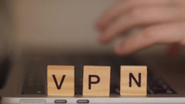 Close-up of wooden letters spelling VPN with hands typing on computer keyboard in background, shallow depth of field
