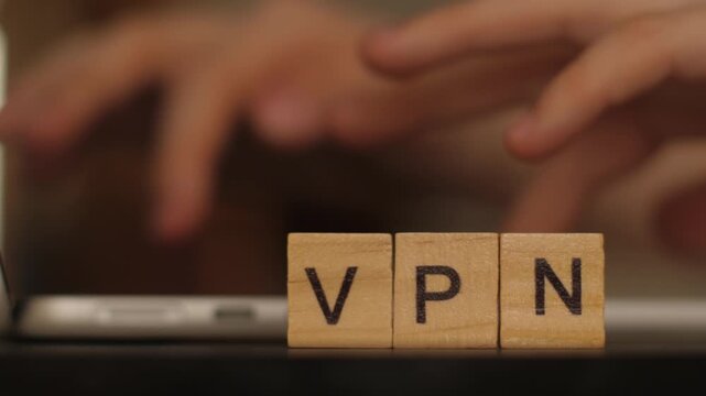 Close-up of wooden letters spelling VPN with hands typing on computer keyboard in background, shallow depth of field
