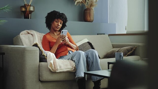 Thoughtful African American woman relaxing on a couch at home scrolling on mobile phone app