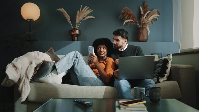 Young multiracial couple relaxing on a couch at home using laptop and mobile phone sharing good relaxing times together