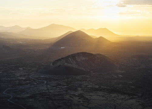 Aerial view of a volcanic landscape bathed in golden light, with the sun's rays piercing through the haze, creating a dramatic contrast between light and shadow, Tinajo, Canary Islands, Spain.