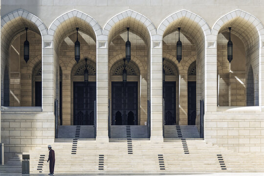 View of grand arches framing dark doorways with hanging lamps, as a man in traditional dress adds a human touch in rd@ lbws`yd, Ad Dakhiliyah Governorate, Oman.