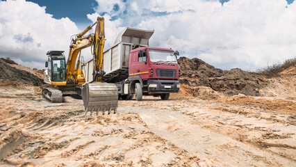 Obraz premium Heavy equipment, including a dump truck and an excavator, works on a construction site with earthen embankments under partly cloudy skies