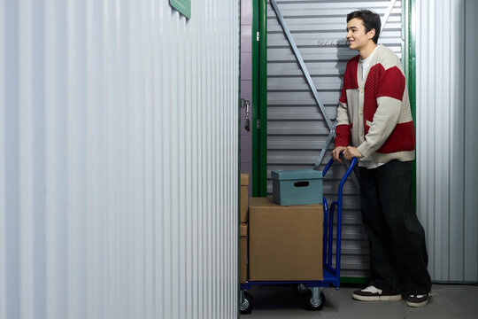 Young adult Caucasian man pushing cart loaded with cardboard boxes and storage container inside self storage facility, standing near metal storage unit door, smiling and looking forward