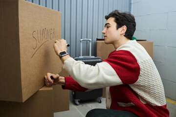 Teenage Caucasian boy labeling cardboard box with marker while organizing storage unit, suitcase and other packed boxes surrounding subject, focused expression on face