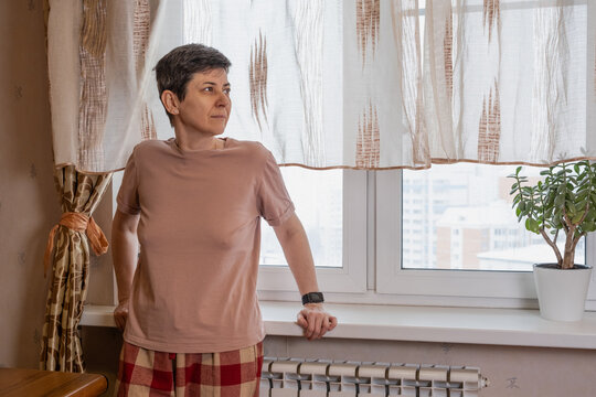 Middle-aged woman leans on a windowsill and looks outside through sheer curtains in a modest apartment, soft daylight interior scene conveying thoughtfulness, home isolation and quiet daytime pause.