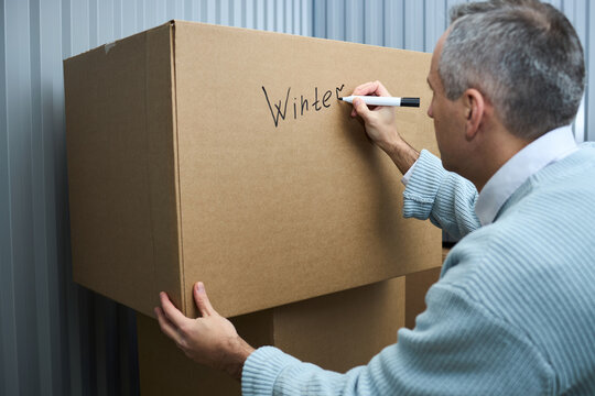 Middle aged Caucasian man labeling large cardboard box with marker, writing word Winter on surface while organizing or preparing items for storage in self storage facility
