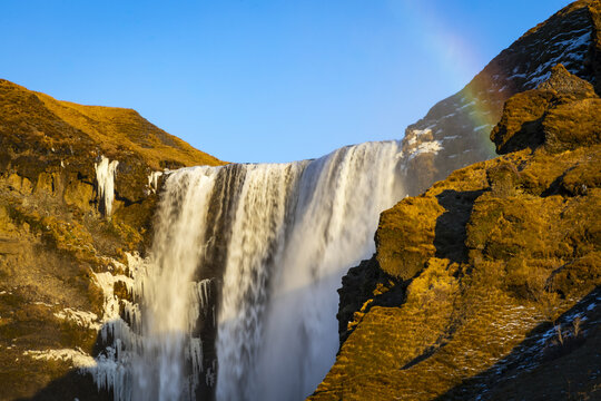 View of the majestic Skogafoss waterfall cascading powerfully from rugged cliffs under a clear sky, a vibrant rainbow arc adding magic, Skogafoss Waterfall, Rangarthing eystra, Iceland.